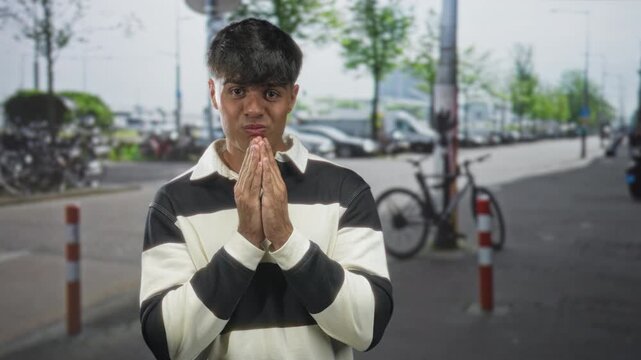 Man with hands clasped praying on a city street sidewalk by bicycles and bollards, face open in surprise and pleading expression; plea hope.