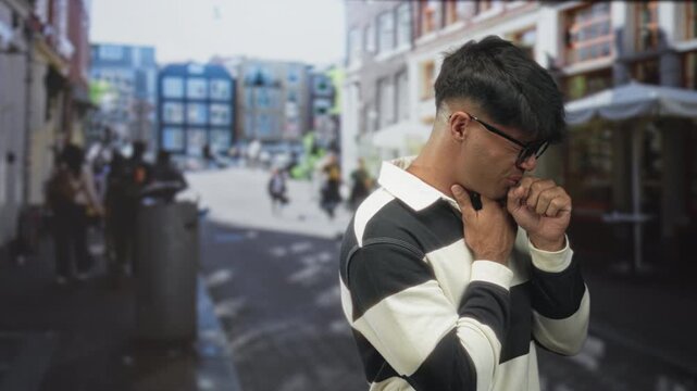 Young man holding throat and coughing into fist on a crowded city street with cafes and pedestrians; discomfort vulnerability sickness.