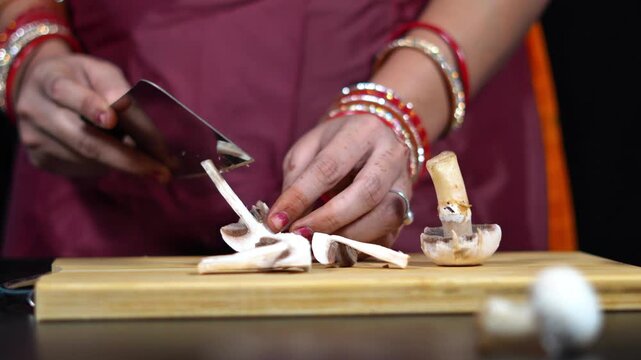 Slicing raw button mushroom with sharp knife on kitchen cutting board. Home cook dicing edible white mushrooms on wooden block in domestic kitchen.