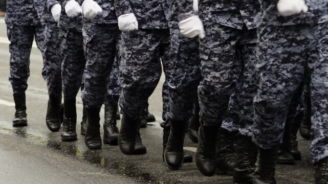 Close-up view of uniformed personnel marching in synchronized formation on a wet street, showcasing black combat boots and digital camouflage trousers during a parade drill