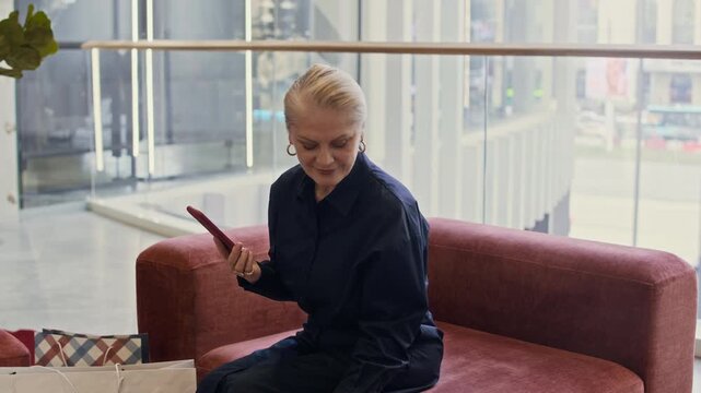 Zoom in slowmo shot of mature Caucasian woman with smartphone in hands checking shopping bags while sitting in mall lounge, reviewing purchase after shopping spree