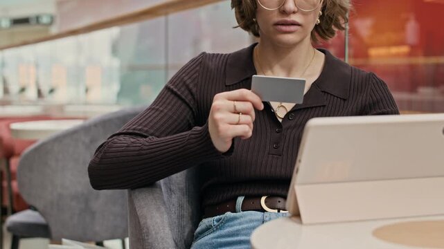 Cropped slowmo midsection shot of young girl in eyeglasses sitting at table in modern mall setting in front of tablet computer and inserting credit card numbers while making online payment