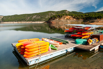 Colorful kayaks and canoes stacked on floating dock at calm reservoir surrounded by pine forest and red rock hills, inland Spain © Bruno Almela