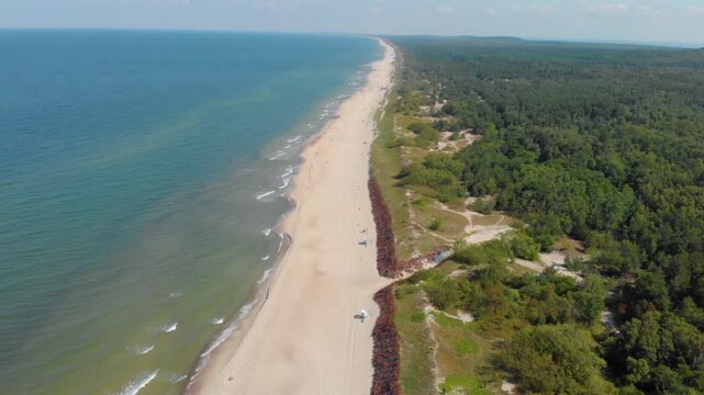 An aerial drone shot on a sunny summer day, looking down the extensive sandy beach of the Baltic Sea, bordered by lush green pine forests and coastal dunes in Palanga, Lithuania.