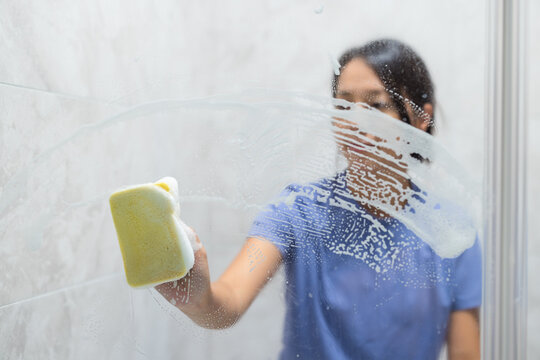 Asian woman wiping soapy bathroom glass door with yellow sponge smiling