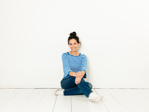 Beautiful young woman with black hair and blue white striped sweater sitting on the ground in front of white background