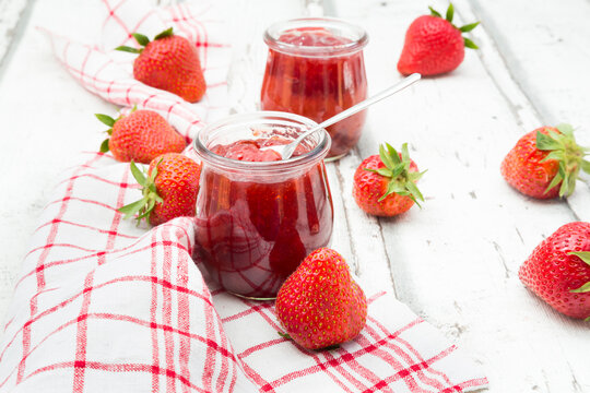 Glass of homemade strawberry jam, kitchen towel and strawberries on white wood