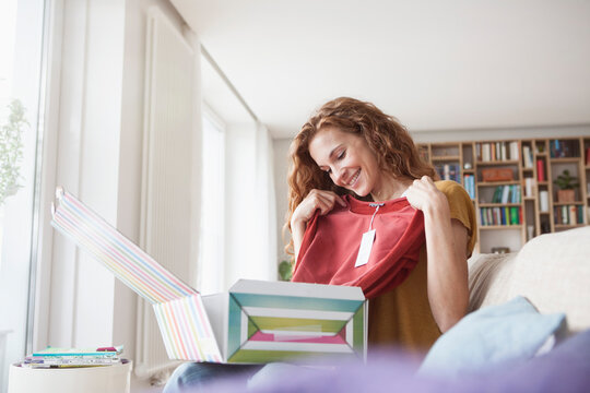 Smiling woman at home sitting on couch unpacking parcel with garment