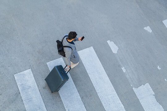 Businessman with baggage crossing the street while looking at cell phone, top view