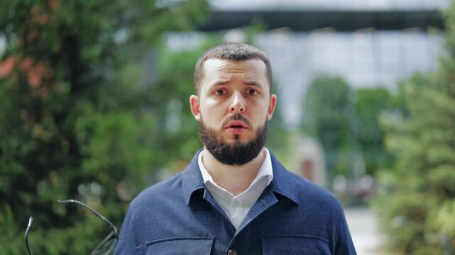 Young bearded man with confused facial expression looking into camera outdoors. Person feels puzzled by strange news, bad offer or technical failure. Great for communication and problem themes.