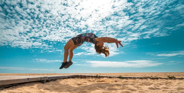 Athletic woman performing a backflip in the air over a sandy path on the beach with the sun shining behind her.