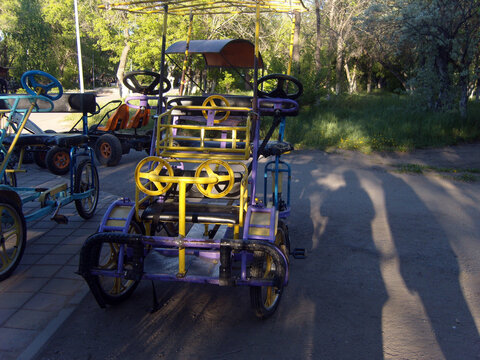 Purple and yellow multi-passenger pedal car parked on a path in a sunny park. Other rental surrey bikes stand nearby under the trees, with long human shadows cast on the ground