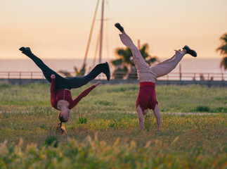 Two athletic women performing impressive handstands and acrobatic flips in a green field during a golden hour sunset © sirbouman