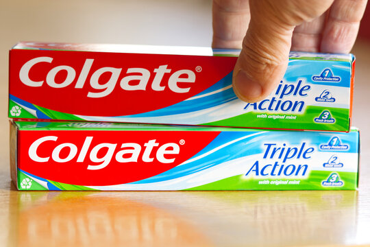 Hand holding one of the two boxes of Colgate toothpaste lying on a table. Colgate is a global oral care brand owned by Colgate-Palmolive, an American consumer goods company.