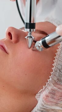 Adult woman receives precise under eye face cheek facial treatment in professional cosmetology clinic. Girl lies relaxed while cosmetologist uses microcurrent tools to rejuvenate skin boost tone.