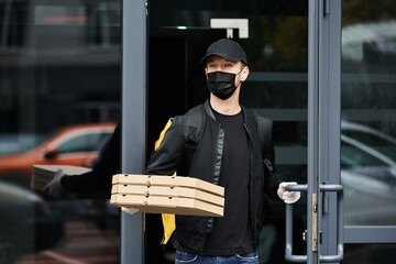 Obraz premium Young man delivers fresh pizza boxes while wearing gloves and a mask in a bustling city