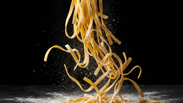 Fresh pasta tagliatelle falling with flour dust against a dark background, ready for cooking.