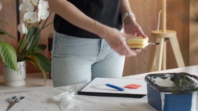 Woman arranges sponge, brush and palette knife on canvas at a covered work table. Concept of DIY art setup, textured painting and handmade decor