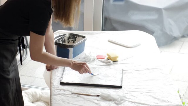Woman spreads white plaster paste across canvas with a palette knife at a covered work table at home. Concept of textured canvas art, handmade wall decor and creative hobby.