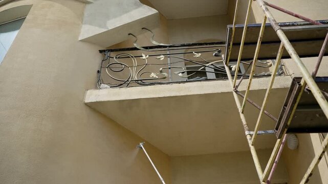 Worker paints building facade with airless spray gun. Professional painter applies beige paint on stucco wall using extension pole. House renovation and exterior improvement process
