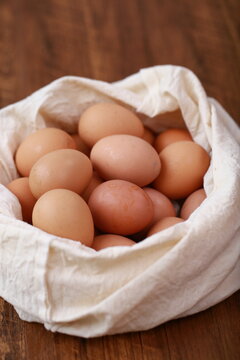 A high-angle, close-up shot shows a cluster of smooth, brown chicken eggs packed inside a cream-colored textile pouch. The bag is gathered at the edges, resting on a warm, dark wood surface. The light