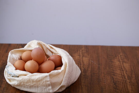 A high-angle, close-up shot shows a cluster of smooth, brown chicken eggs packed inside a cream-colored textile pouch. The bag is gathered at the edges, resting on a warm, dark wood surface. The light