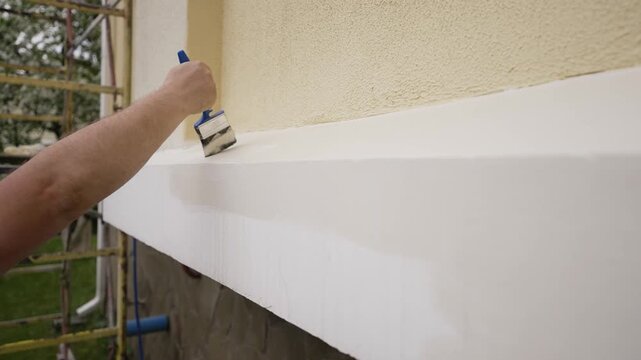 Close up of a painter's hand applying fresh white paint to an exterior house ledge with a brush. Home renovation, construction, and DIY improvement project in progress outdoors