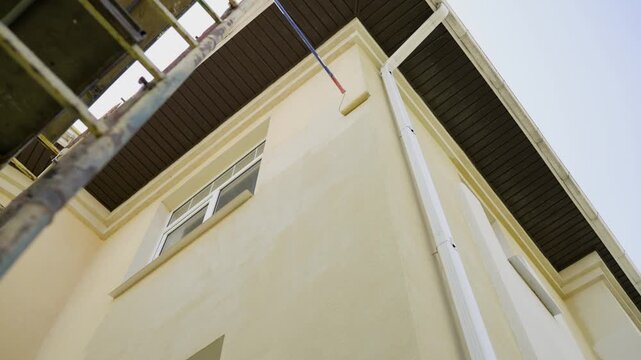 Low angle shot shows a painter applying light yellow paint to a house facade with a roller on an extension pole. Scaffolding in view. Home improvement and renovation in progress