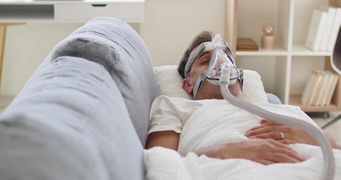 Man with mask sleep apnea therapy oxygen. A male on a couch uses a CPAP device for airway support and treatment at home to improve sleep safety. Concept healthcare breathing therapy for apnea.
