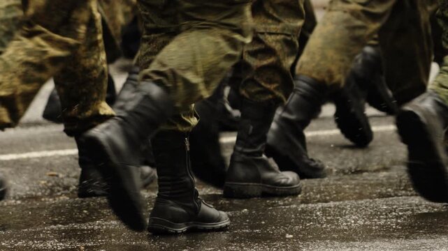 Close-up view of uniformed soldiers in camouflage marching in formation on a wet asphalt road, heavy black combat boots striking the pavement in synchronized steps during drill