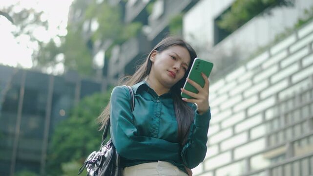 Anxious young Asian businesswoman looking at her smartphone with a worried expression on a city street, feeling stressed by bad news or confused by a message, Anxious, Sad, Wrong, Depressed.