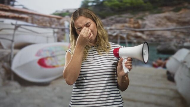 Woman holding megaphone pinches nose from strong odor at rocky beach cove with stacked inflatable boats and dock steps; disgust protest odor.