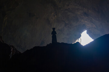 Silhouette stands inside vast cave. Light floods in from distant opening. Cave walls rise steep and dark. Figure appears small against cavern. Natural cavern interior © jinhongljh