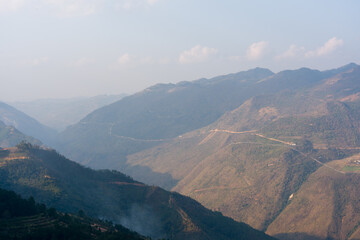 Obraz premium Mountain range stretches across the horizon. Winds stir the dry, sun-baked slopes. Distant ridges fade beneath a hazy sky. Faint trail winds along the valley's edge