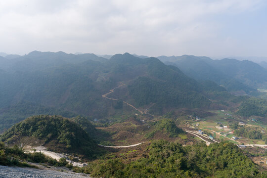 Rolling hills stretch under soft, cloudy sky. Green slopes meet winding rural roads. Village dots distant valleys below. Trees and shrubs cover rugged terrain. Mountains fade into hazy horizon