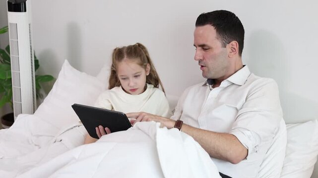 Father and daughter using tablet together while relaxing in bed at home. Family bonding, parenting, online learning, digital lifestyle, and quality time concept.