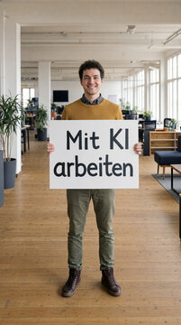 Junger Mann im gelben Pullover mit gro&szlig;em Schild Mit KI arbeiten im gro&szlig;en Loft B&uuml;ro Totale &ndash; K&uuml;nstliche Intelligenz am Arbeitsplatz Wide Shot