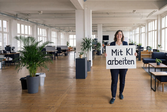Klassisch deutsche Frau &uuml;ber 50 mit gro&szlig;em Schild Mit KI arbeiten im weiten Loft B&uuml;ro &ndash; K&uuml;nstliche Intelligenz am Arbeitsplatz Wide Shot Establishing