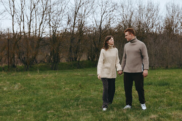 Fototapeta premium A couple walks hand in hand in a grassy park, wearing cozy sweaters, enjoying a calm outdoors moment in autumn landscape, warm mood and simple lifestyle.