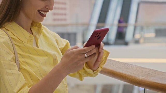 Cropped slowmo shot of blonde woman in yellow shirt smiling while texting message on smartphone, standing by handrail in shopping mall with escalator moving in blurred background