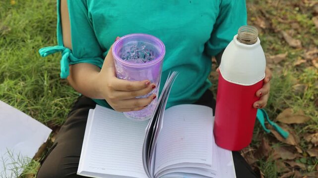  Young Indian girl sitting in a green park while pouring water from a bottle into a sipper and keeping a book on her lap during hot summer weather. Outdoor study and hydration lifestyle mo
