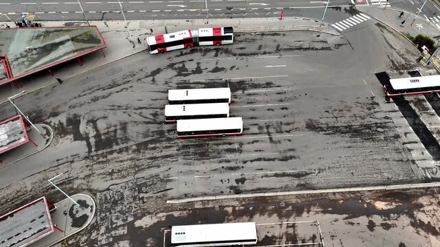 High-angle aerial drone footage of a bustling urban bus terminal with multiple buses maneuvering and vehicle traffic on an adjacent city street, symbolizing public transit and infrastructure.