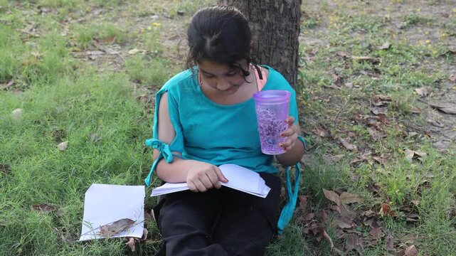 Young Indian girl sitting under a tree in a green park while reading a book and holding a water bottle during hot summer weather. Peaceful outdoor study and hydration lifestyle moment in India.