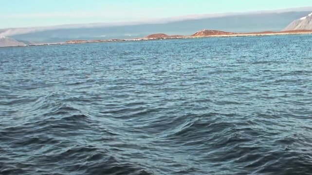Marvel at the endless, deep blue of the Arctic Ocean, where serene waves dance under a bright sky. A distant, rugged landmass graces the horizon, marking a passage through this profound polar realm.