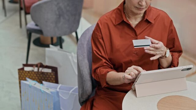 High angle cropped slowmo shot of elegant adult woman sitting at table in mall among shopping bags and inserting credit card numbers while making online payment on digital tablet