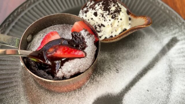 Close-up of a silver spoon breaking into a warm chocolate lava cake with a molten center, served with vanilla ice cream and fresh strawberries.