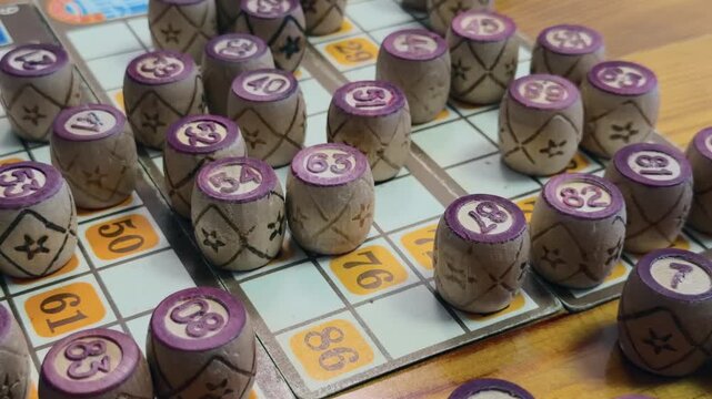 Wooden Bingo Barrels Standing on Vintage Lottery Cards and Rustic Wooden Table as Camera Pulls Back from Close Up to Wide View Revealing Full Board Game Layout in Warm Indoor Lighting Cozy Atmosphere