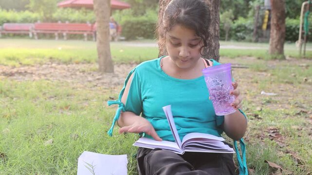  Young Indian girl sitting peacefully in a green park while reading a book and holding a water bottle during hot summer weather. Calm outdoor study and hydration lifestyle moment in India.
