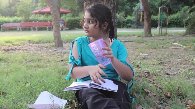  Young Indian girl sitting under a tree in a green park while reading a book and holding a water bottle during hot summer weather. Peaceful outdoor study and hydration moment in India.