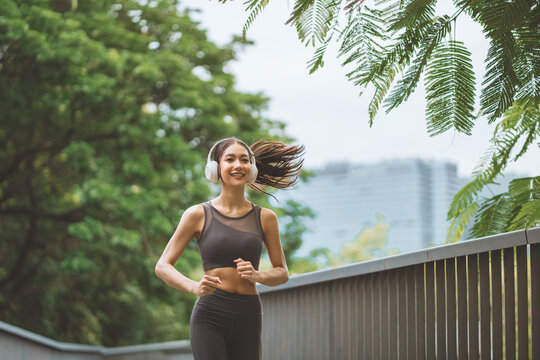 Cheerful young Asian woman running at public park bridge wearing headphones listening to music happy active female jogging for morning workout urban fitness lifestyle and wellness routine concept